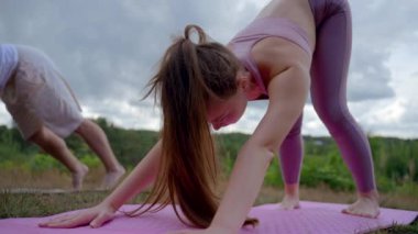 Young woman does yoga pose on mat on green hill. Athletic lady in sportswear concentrates on asana Upward-Facing Dog against cloudy sky low angle shot
