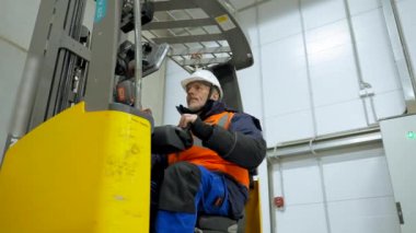 Engineer controls forklift truck pushing buttons on monitor. Bearded man sits in cabin of machine in storage terminal low angle shot slow motion