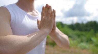 Young muscular man meditates in Lotus pose folding hands in namaste on high grass hill. Sporty guy enjoys practicing yoga sitting on mat against green trees