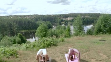 Young people practice yoga asana together on mats on grassy hill. Concentrated man and woman perform Wheel pose and unite with nature near river upper view