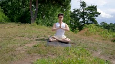 Young athletic man meditates in Lotus pose with namaste closing eyes on grassy meadow. Guy in comfortable clothes sits on mat breathing fresh air closeup