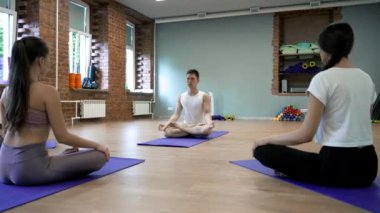 Male coach conducts yoga class for young women in gym. People with closed eyes sit in Lotus pose on blue mats put on wooden floor in light studio
