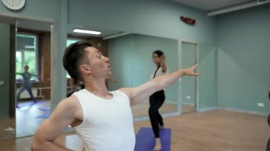 Professional male coach shows yoga exercise to women students standing on blue mats. Young man and ladies enjoy training in big hall closeup