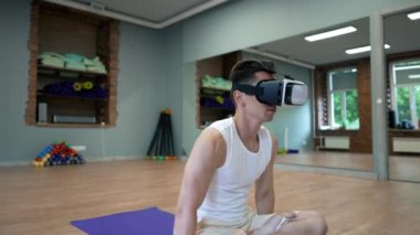 Young man in VR goggles enjoys doing yoga exercise on mat put on wooden floor in big hall. Male person takes care of body and health
