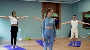 Kadın, Asana 'yı hasırların üzerinde yapanlara karşı kameraya dönüyor. Genç koç yoga sınıfında stüdyoda durup gülümsüyor. Ağır çekim yakın çekim.