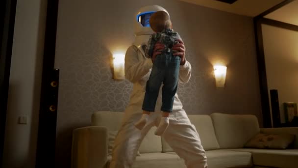 一位身穿航天服的男性父亲开始与蹒跚学步的儿子在舒适的公寓里跳舞 让孩子们在宇宙飞船上开始他有趣的太空工作的概念 — 图库视频影像