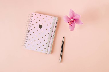 Kalpli bir defter ve pembe arkaplanda pembe bir orkide ile duran bir kalem. Notlar, düşünceler, planlar, günlük ...