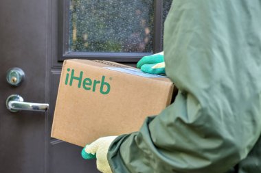 iHerb logo on a cardboard box in the hands of the courier during delivery to the door of the house: Riga, Latvia - September 10, 2021