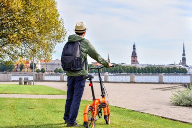 Letonya 'nın Riga kentinin arka planına karşı Daugava Nehri yakınındaki bir şehir parkında bisikletli bir adam duruyor..