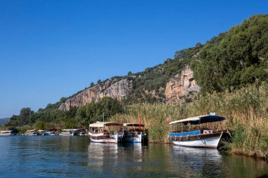 Dalyan, Mugla, Türkiye - 22 Ekim 2021: Dalyan, Mugla, Türkiye 'deki Kaunos antik kentinin kaya kesimli tapınak mezarları. Feribot ve arka planda oyulmuş mezarlarla güzel Dalyan nehir manzarası.