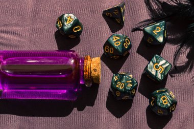 Close-up image of a set of dark role-playing gaming dice and a glass bottle with pink liquid in the sun
