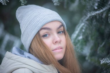 Karlı bir parkta gri şapkalı bir sarışın, donmuş çam dallarının arkasında bir kadının portresi, kış ormanındaki güzel genç bir kadın.