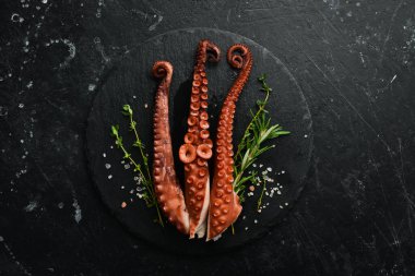 Metal bir tabakta baharat ve otlarla haşlanmış ahtapot dokunaçları. Siyah taştan bir arka planda.