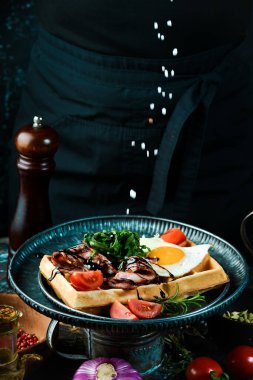 Yumurtalı sandviç, domates ve domuz pastırması bir şefin elinde, siyah bir arka planda. Belçika Waffle 'ları.