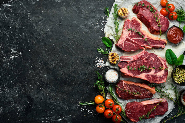 Variety of raw aged beef steaks: t-bone, tomahawk, striploin, tenderloin, new york steak for grilling with spices on stone background. On a black stone background.