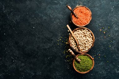 Kâselerdeki organik tahıllar: nohut, fasulye, mercimek. Sağlıklı yiyecekler. Siyah taştan bir arka planda.