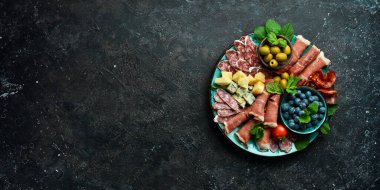 İtalyan atıştırmalıkları. Peynirli, jambonlu, jambonlu, jambonlu ve abur cuburlu tabak. Siyah taştan bir arka planda.