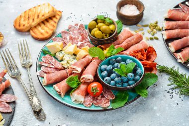 Farklı çeşit peynir ve jambon, prosciutto, jamon salamı ve aperatiflerle dolu bir tabak. Antipasto ya da aperatif parti konsepti. Gri taştan bir arka planda.