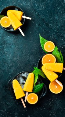 Çubukta portakallı dondurma. Taze portakallar ve sarı buzlu şekerler. Siyah taştan bir arka planda. Üst görünüm.