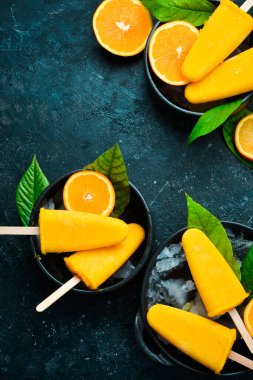 Çubukta portakallı dondurma. Taze portakallar ve sarı buzlu şekerler. Siyah taştan bir arka planda. Üst görünüm.