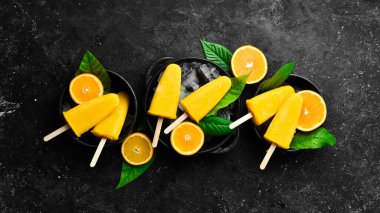 Çubukta portakallı dondurma. Taze portakallar ve sarı buzlu şekerler. Siyah taştan bir arka planda. Üst görünüm.