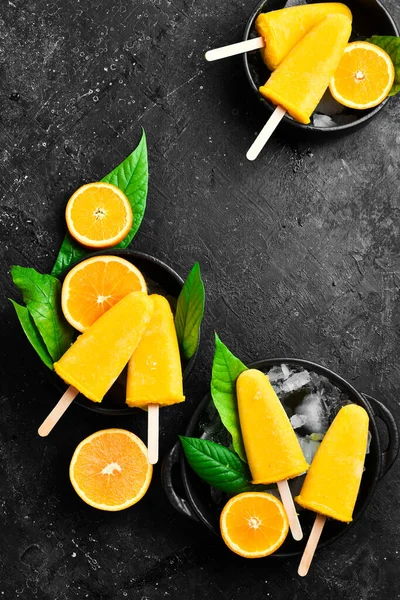 Orange ice cream on a stick. Fresh oranges and yellow popsicles. On a black stone background. Top view.