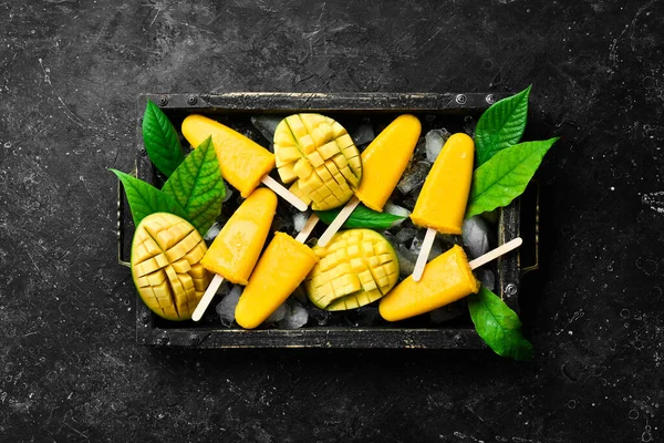 Frozen mango popsicles on a tray. Cold summer sweets. On a black stone background. Top view.