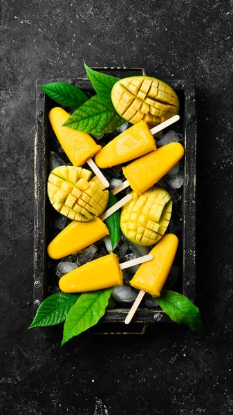 Frozen mango popsicles on a tray. Cold summer sweets. On a black stone background. Top view.