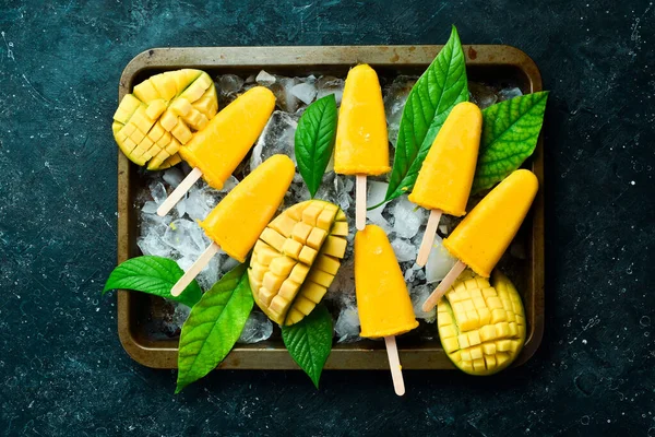 Frozen mango popsicles on a tray. Cold summer sweets. On a black stone background. Top view.