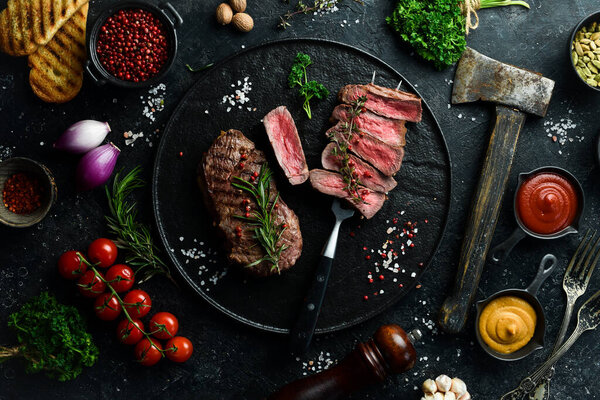 Grilled ribeye beef steak. Steak on a fork on a black plate. Top view. On a black stone background.