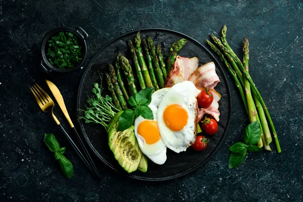 Tasty breakfast. Sunny side up eggs, asparagus, bacon and avocado. On a black stone plate.