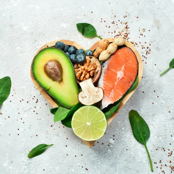 Foods good for the heart: nuts, salmon, avocados, spinach, mushrooms, berries. In a heart-shaped box. On a stone background. Top view. Copy space.