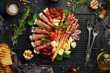 İtalyan malzemeleri: jambon, jambon, salam, parmesan, zeytin, ekmek çubukları. Siyah taştan bir arka planda.