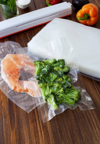 Fresh pieces of broccoli in a vacuum bag and a piece of red fish next to a pack of vacuum bags and a vacuum cleaner on a dark wooden table. Copy space text