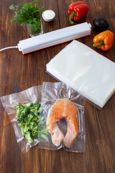 A piece of salmon in a vacuum bag and broccoli next to a pack of vacuum bags and a vacuum cleaner on a dark wooden table. Copy space text