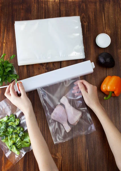 The process of sealing a vacuum bag with chicken legs with a vacuum cleaner next to packed vegetables on a wooden dark table. Copy space text