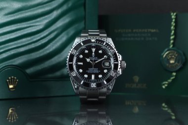 Rolex klasik kol saati seramik model siyah istiridye daimi denizaltı tarihi ayna rafında arka planda Rolex Green Crown logosu, bozuk para ve el kitabı ile 39 mm.