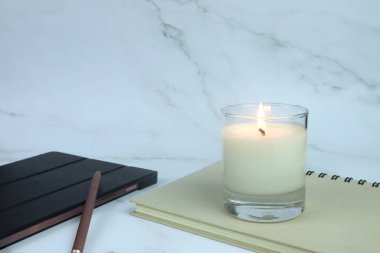 Beyaz mermer masada beyaz mermer mum kokulu bir bardak aroma ve beyaz yatak odasında bir tablet var.