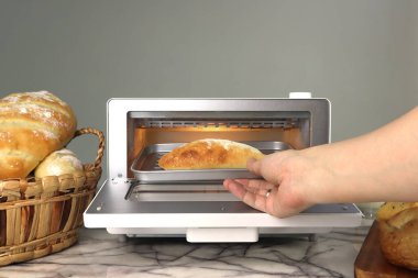 Bayan bir ev kadınının sağ eli ekmeği beyaz modern tasarım tost makinesinin içine koydu. Ekmek mermer masanın üzerinde, mutfak odasının arka planındaki gri beton duvarda ev yapımı tost ekmeği vardı.