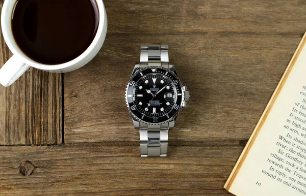 Lüks Rolex klasik kol saati seramik model siyah istiridye daimi denizaltı tarihli 44 mm, sabah kahvaltısı sırasında bir kitap ve bir fincan kahveyle ahşap masaya konur.