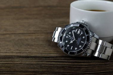Lüks Rolex klasik kol saati seramik model siyah istiridye daimi denizaltı tarihli 40 mm, sabah kahvaltısı sırasında bir kitap ve bir fincan kahveyle ahşap masaya konur.