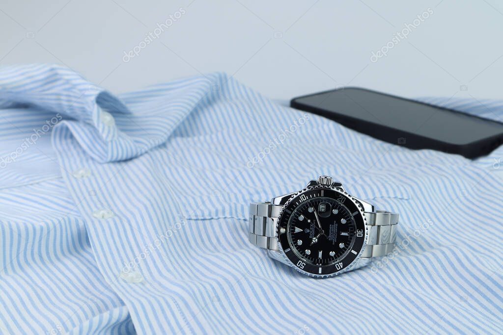 Luxurious Rolex vintage wristwatch ceramic bezel model black oyster perpetual submariner date 40 mm is put on white wooden table with blue shirt and mobile phone in the morning before working time