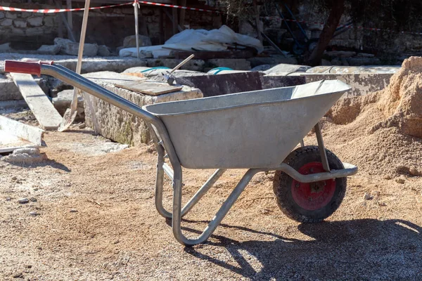 Empty metal wheelbarrow, shovel and other tools at excavation site in Side, Turkey. Ancient ruins in background on sunny day. Selective focus.
