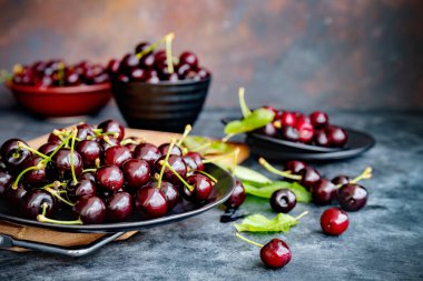 Koyu arkaplanda gri seramik tabakta yeşil yapraklı taze siyah kiraz. Karanlık, sade bir fotoğraf. Seçici odaklanma. Bulanık arkaplan.