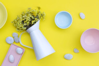 Bouquet of dried Helichrysum flowers in white ceramic jug, blue, yellow and pink ceramic bowls, pink rectangular plate and smooth sea stones on bright yellow background. Top view, flat lay composition.
