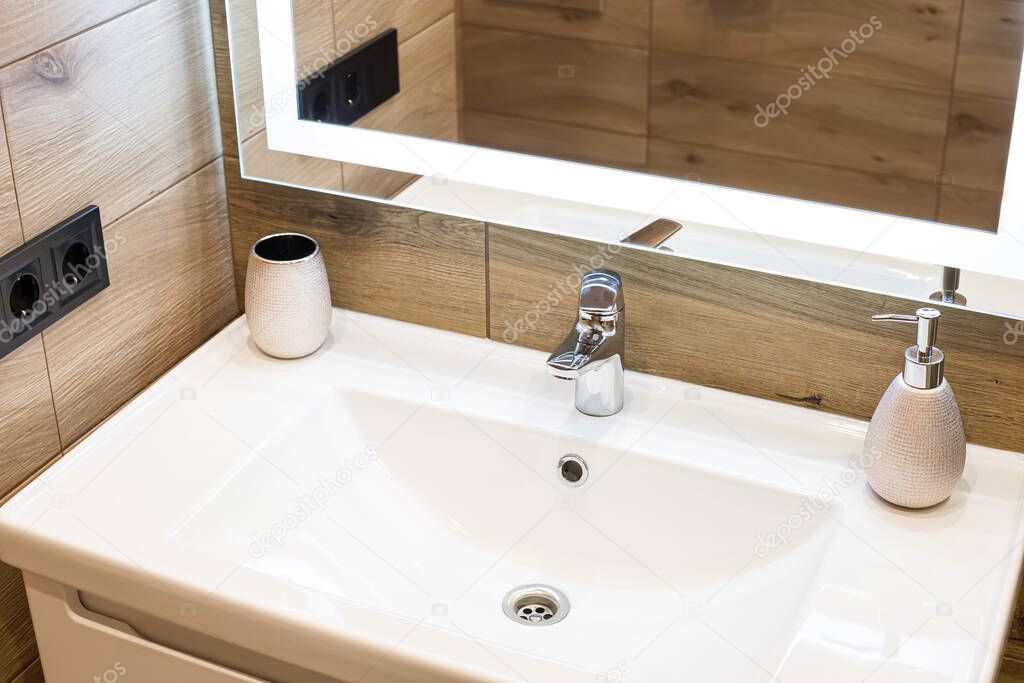 Modern washbasin with chrome faucet beside a stylish soap dispenser. Mirror with built-in led lighting.
