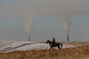 Almaty, Kazakistan, 12.01.2021. Ata binen bir binici bozkırda bir kömür santralinin bacalarının arka planına çarpar.