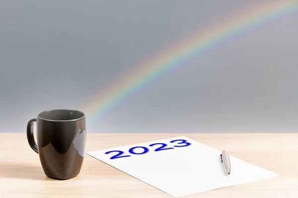 Number 2023 written on a paper, with a mug, a pen and a rainbow background. New year concept. invitation