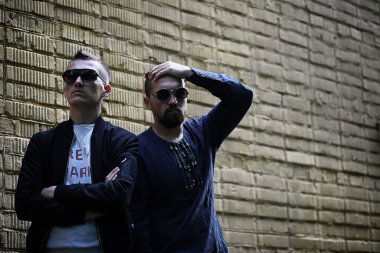 Güneş gözlüklü iki adam tuğla duvara yaslanmış.