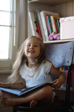 Küçük kız odada oturuyor ve kitap okuyor.